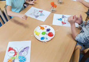 Dzieci przy stoliku ozdabiają wizytówkę patyczkami kosmetycznymi umoczonymi w kolorowej farbie