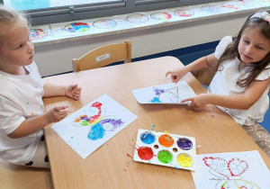 Dzieci przy stoliku ozdabiają wizytówkę patyczkami kosmetycznymi umoczonymi w kolorowej farbie