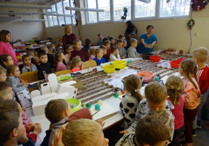 Dzieci stoją wokół stołu warsztatowego, pani tłumaczy im co będą robiły