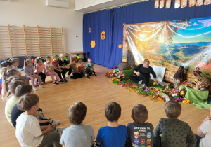 Dzieci siedzą w kręgu przed panem, który pokazuje przedmioty dotyczące życia mrówek