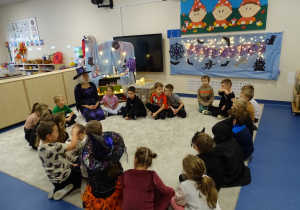 Dzieci z panią w stroju czarodziejki siedzą w kole w sali ustrojonej na andrzejki