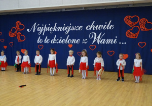 Grupa dzieci stoi na scenie podczas występu, w tle dekoracja z czerwonymi sercami i napisem.