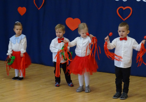 Kilkoro dzieci w strojach galowych gra na prostych instrumentach perkusyjnych.