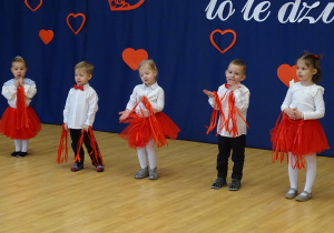Chłopiec trzyma mikrofon, obok stoją inne dzieci z rekwizytami.