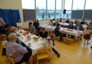 Goście siedzą przy stołach podczas spotkania w sali przedszkolnej.
